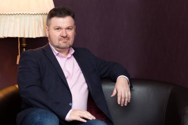 Kanepede oturan bir ceketli bir sakallı sağlam adam.