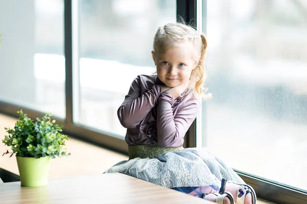 4-5 yaşında beyaz saçlı bir kız, bir kafede bir pencerenin yakınında oturuyor. Küçük bir sarışın kız büyük pencere tarafından oturuyor.