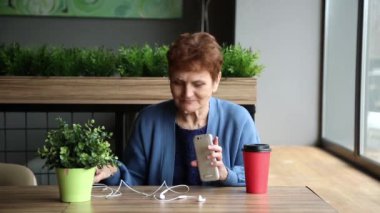 Kulaklık ve telefon ile siğil yaşlı kadın. Telefon ve bir kafede bir smartphone kulaklık ile kadın emeklinin. Emeklici kulaklarına kulaklık ekler. Papilloma siğiller üzerinde kadın yüz.