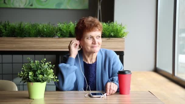ヘッドセットと電話でいぼを持つ年配の女性 女性は携帯電話からのヘッドホンとカフェのスマートフォンで年金受給者 年金受給者は彼女の耳にヘッドホンを挿入します 女性の顔の乳頭腫疣贅 — ストック動画