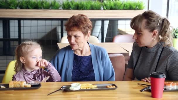 小さな女の子は彼女の母親と祖母と話していますカフェで子供が彼の祖母と母にその日のニュースを伝えます — ストック動画