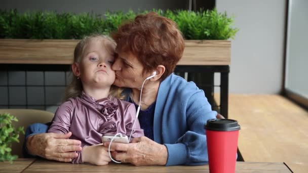 孫娘と一緒に祖母は電話のヘッドフォンで音楽を聴きます 高齢の退職した女性と小さな女の子が スマートフォンのヘッドフォンで歌を聴きながらカフェに座っています ファミリ値 — ストック動画