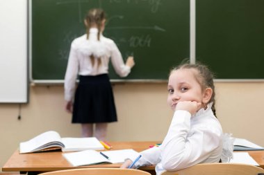 Bir arkadaşı tahtaya yazarken liseli kız geriye bakıyor. Bir kız öğrenci kız kameraya bakarken arkadaşı, bir sınıf arkadaşı, tahtadaki öğretmene cevap verir..