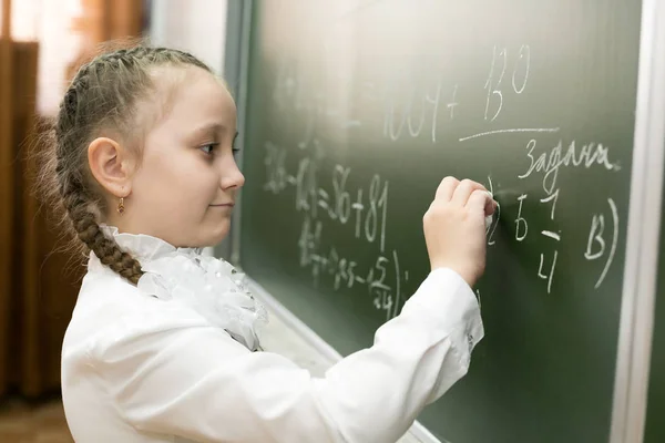 女子小学生が黒板に書き込む 小さな女子高生は 彼女の手にチョークを保持し 学校の黒板に問題の解決策を書きます — ストック写真