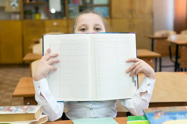 The schoolgirl covered her face with a school gradebook. A teenager girl hides her face behind an open book raised by a school book.