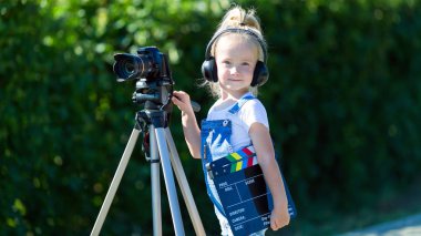 Bir tripod üzerinde bir kamera ile küçük bir kız kulaklık bir şehir parkı duruyor. Bir kamera ve bir tripod ile Çocuk acemi video blogger.