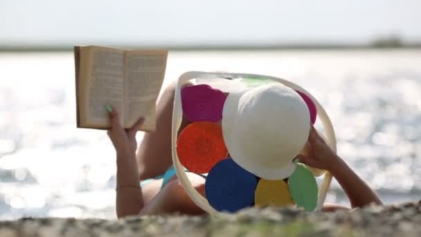 その少女は本を持って浜辺に横たわっている 水着と帽子をかぶった若い女性がビーチで日光浴をし 背中に横たわり 本を読んでいる — ストック動画