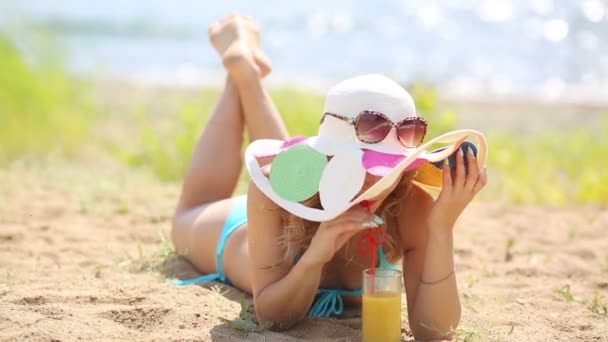 帽子をかぶった若い女性は海岸で休息を取り 彼女の胃の上に横たまり オレンジジュースを飲みます — ストック動画
