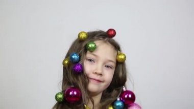 Kafasında Noel süsleri olan bir kız. Küçük bir kız saçlarında ağaç süslemek için Noel topları örgülü, elinde iki güzel topları tutar.