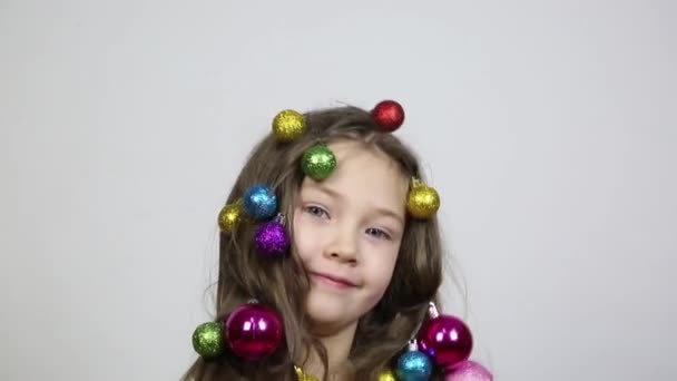 彼女の頭の上にクリスマスの装飾を持つ女の子 小さな女の子は 彼女の髪に木を飾るためにクリスマスボールを編組し 彼女の手に2つの美しいボールを保持しています — ストック動画