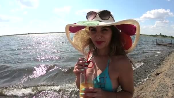 帽子をかぶった少女は水のところでジュースを飲む 帽子をかぶった若い女性が川沿いのビーチでオレンジジュースを飲む — ストック動画