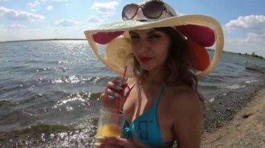 Şapkalı bir kadın sahilde yatıyor ve sahilde bir bardak meyve suyu ile ananas suyu Kız içiyor 