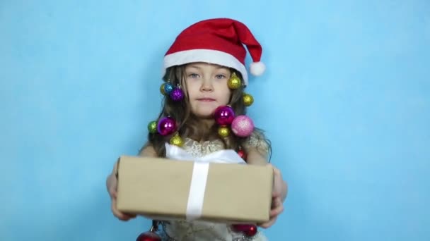 クリスマスの帽子の子供が手に贈り物を持っている 彼女の髪にクリスマスボールと美しいドレスの女の子は彼女の手の中にクリスマスのための驚きのギフトボックスを持っています — ストック動画