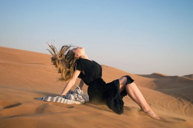 The girl develops hair from the wind in the desert. A young girl in a black dress sits on the sand in a beautiful pose with her hair developing in the wind. clipart