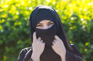 Yeşil yapraklı güzel bir müslüman kadın. Doğulu giysiler içindeki İslami kadın, yoğun yeşil çalıların arkasında duruyor..