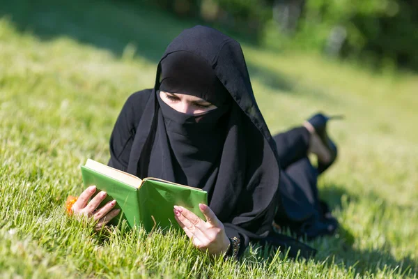 イスラム教徒の女性は公園の芝生の上で本を読みます イスラム教徒の学生の女の子は 試験のために準備草の上に横たわる教科書を読みます — ストック写真