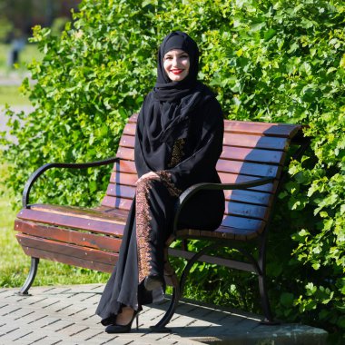 Genç ve güzel bir müslüman kadın bankta oturuyor. İslami kız, bir bankta dinlenirken geniş bir gülümseme sergiliyor.