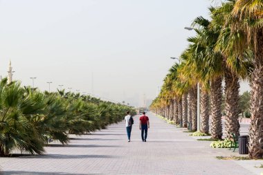 Deniz kıyısı boyunca geniş yaya yolu. İnsanlar, palmiye ağaçları ve tropikal bitkilerin yetiştiği geniş kaldırımda yürürler..