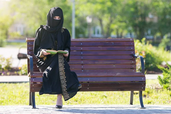 身穿黑色民族服装的阿拉伯妇女正在一个城市公园看书 一位来自中东的移民正坐在长椅上 — 图库照片