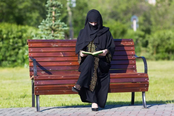 A migrant from the Middle East is sitting on a bench.  Arab woman in black national dress is reading a book in a city park