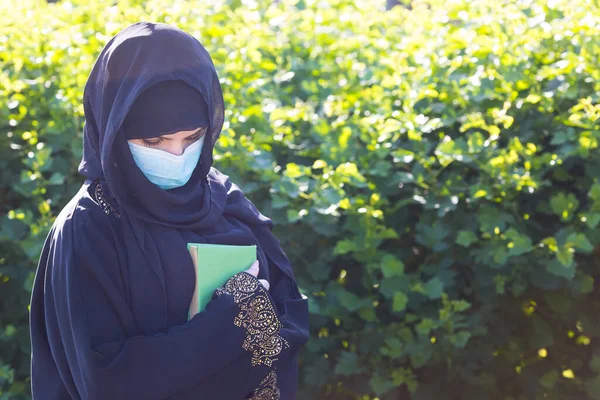 A migrant from the in a mask on her face and national clothes holds in her hands a green Karan against a background of trees. Oriental woman in a protective medical mask quaran in her hands.