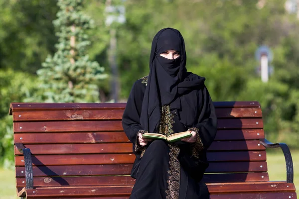 身穿黑色民族服装的阿拉伯妇女正在城市公园看书 一位来自中东的移民正坐在长椅上 — 图库照片