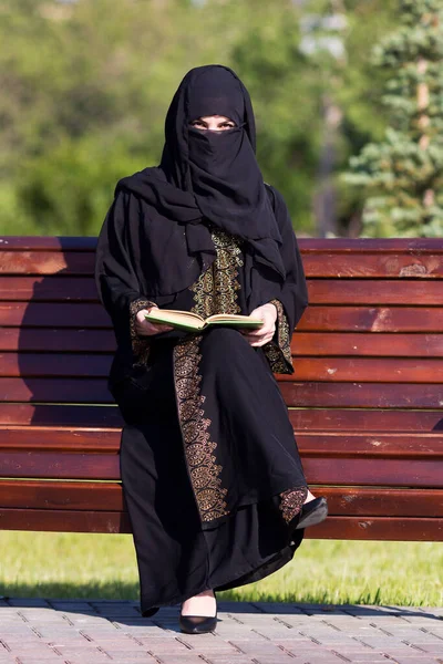 A migrant from the Middle East is sitting on a bench. Arab woman black national dress is reading a book in a city park.