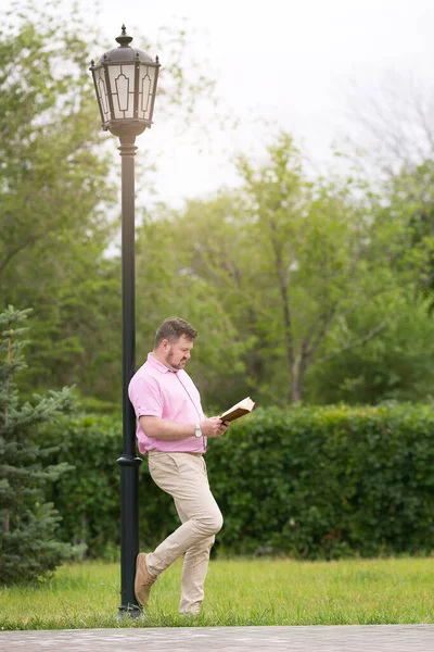 A man in a pink shirt with a book in his hands stands near a lighting pole in a park.