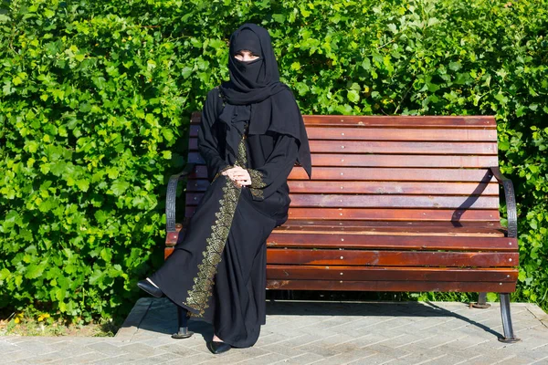 Arab woman migrant from the Middle East in black national clothes is sitting on a bench. in a city park.