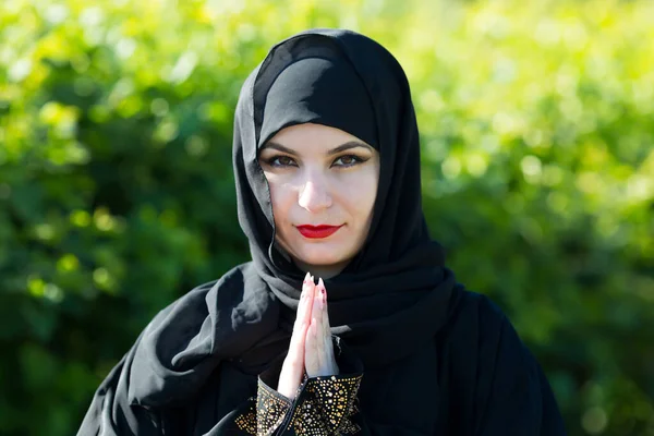 Arab woman in black clothes prays to God against a background of green trees