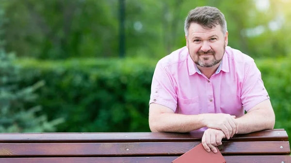 A grown man with a beard in a red shirt is leaning against a bench in the park. He holds a book in his hands.
