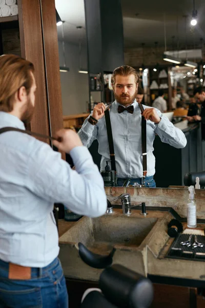 Man Dressing Up Near Mirror In Salon. Portrait Of Handsome Male Barber Wearing Suspenders. High Resolution