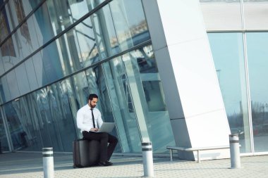 İş adamı havaalanında seyahat bilgisayar üzerinde çalışıyor. Bagaj açık havada oturup dizüstü kullanarak bavul olan adam. Yüksek çözünürlük