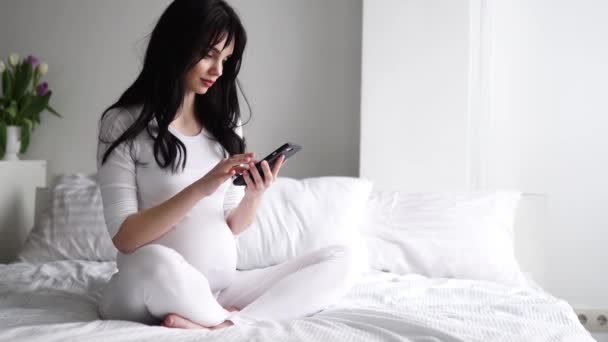 Terhes nő mobiltelefonnal ül az ágyon a hálószobában — Stock videók