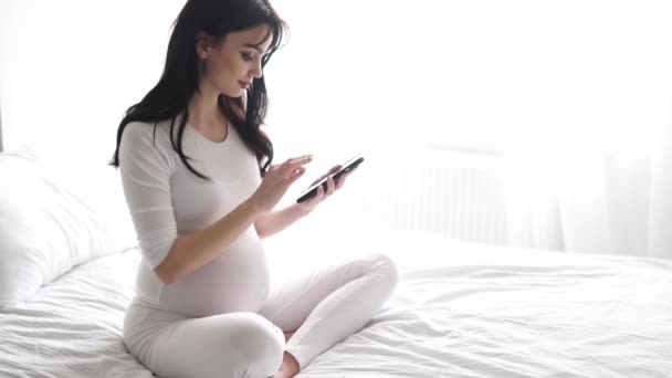 Gravid kvinna med mobiltelefon sitter på sängen i sovrummet — Stockvideo