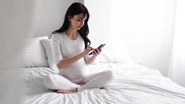 Mulher grávida com telefone móvel sentado na cama no quarto — Vídeo de Stock