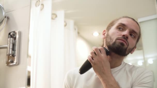 Мужчина бреется лицом с триммером, смотрящим в зеркало — стоковое видео