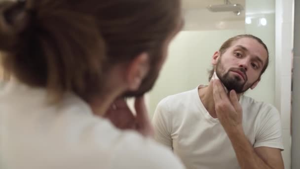 Hombre aplicando espuma de afeitar en la piel de la cara y mirando en el espejo — Vídeo de stock