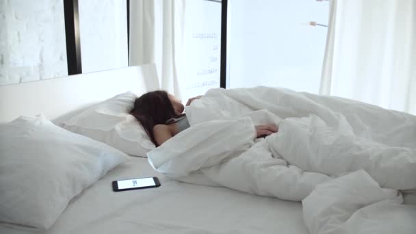 Будильник на телефоне. Уставшая женщина просыпается в постели в спальне — стоковое видео