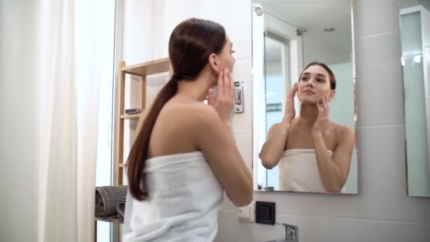 Уход за кожей. Женщина трогает лицо и смотрит в зеркало на ванную — стоковое видео