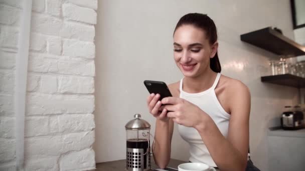 Χαμογελαστή γυναίκα με κινητό τηλέφωνο, πίνοντας τον καφέ στην κουζίνα — Αρχείο Βίντεο