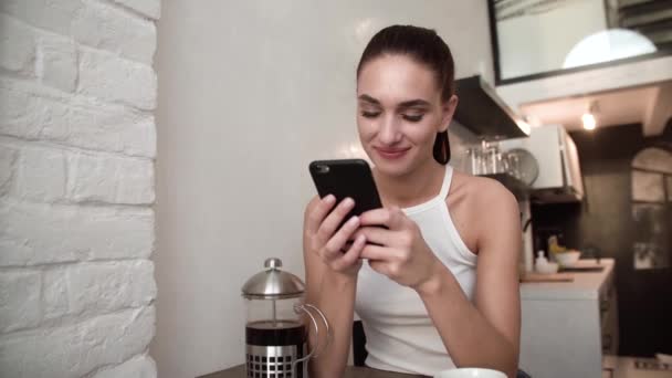 Улыбающаяся женщина с мобильного телефона пьет кофе на кухне — стоковое видео
