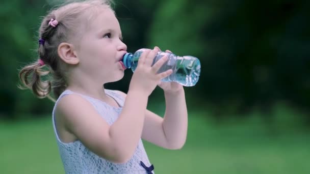 Пейте воду. Маленькая девочка пьет воду из бутылки на открытом воздухе — стоковое видео