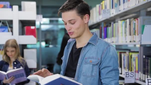 Biblioteca. Sonriente estudiante masculino lectura libro cerca de estantes — Vídeos de Stock