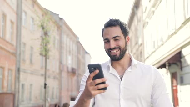 Smiling Man Using Mobile Phone and Walking Along City Street — стоковое видео