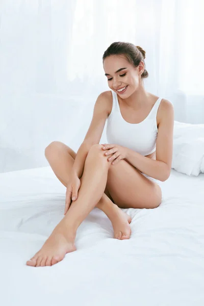 Уход за кожей тела. Женщина, трогающая длинные ноги на белой кровати — стоковое фото