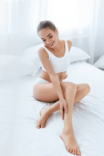 Догляд за шкірою тіла. Красива жінка торкається довгих ніг на білому ліжку — стокове фото