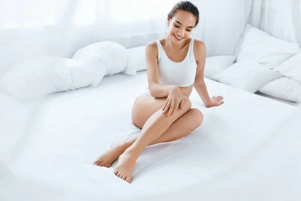 Cuidados com a pele corporal. Mulher bonita tocando pernas longas na cama branca — Fotografia de Stock