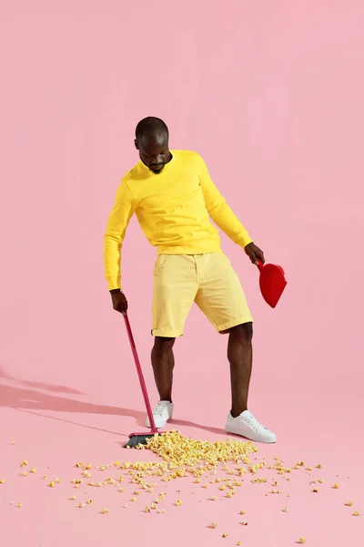 Black man sweeping pop corn on floor on pink background