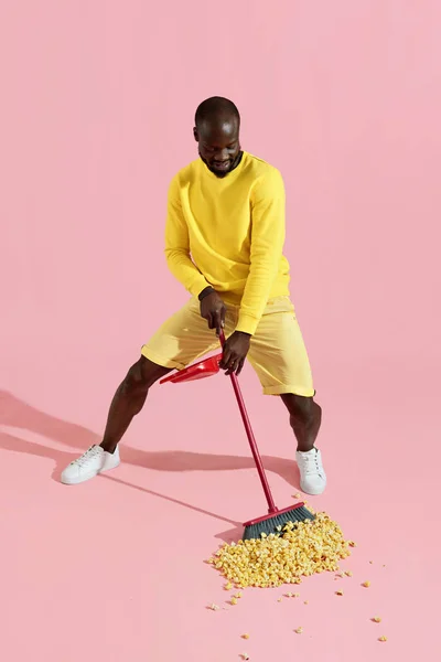 Hombre negro barriendo maíz pop en el suelo sobre fondo rosa — Foto de Stock